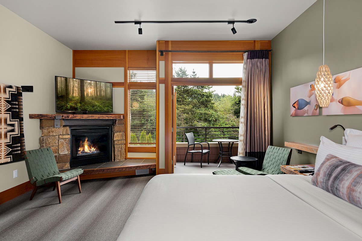 This image shows a hotel room with a bed, fireplace, TV, chairs, and a balcony with a view of trees. Modern decor includes artwork and pendant lighting.