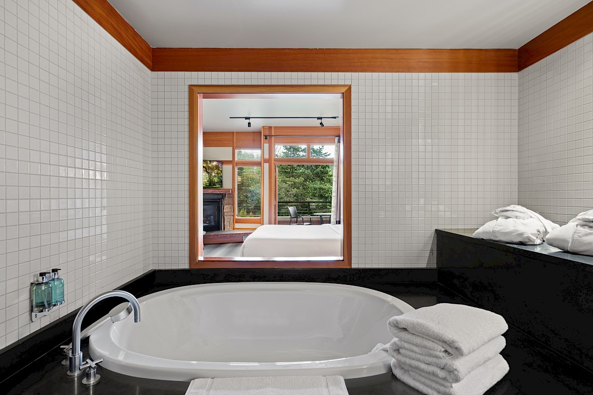 The image shows a bathroom with a round bathtub, towels, and a view into a bedroom through a windowed wall.