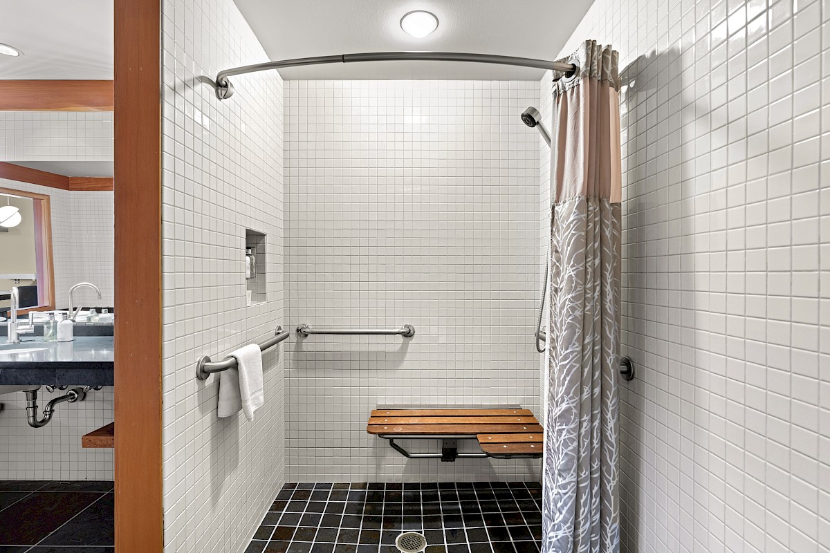 A modern bathroom shower area with white tiles, a bench, safety bars, a shower curtain, and a towel hanging on a rail.