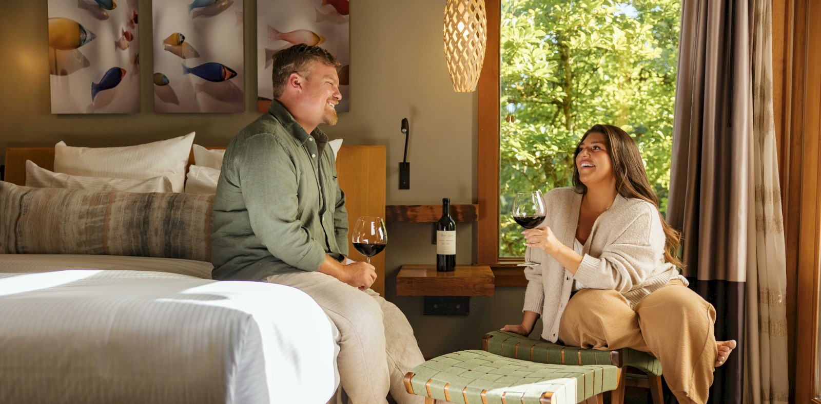 Two people share a cozy hotel room, lounging on a bed and a chair, enjoying wine and conversation near a windowlit scene.