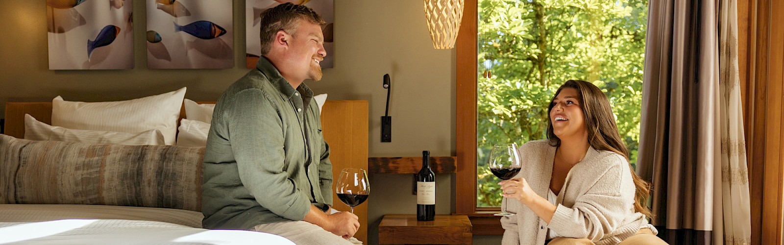 Two people sit in a cozy hotel room, sharing wine by a bed and a small table, enjoying a relaxed chat and a bottle.