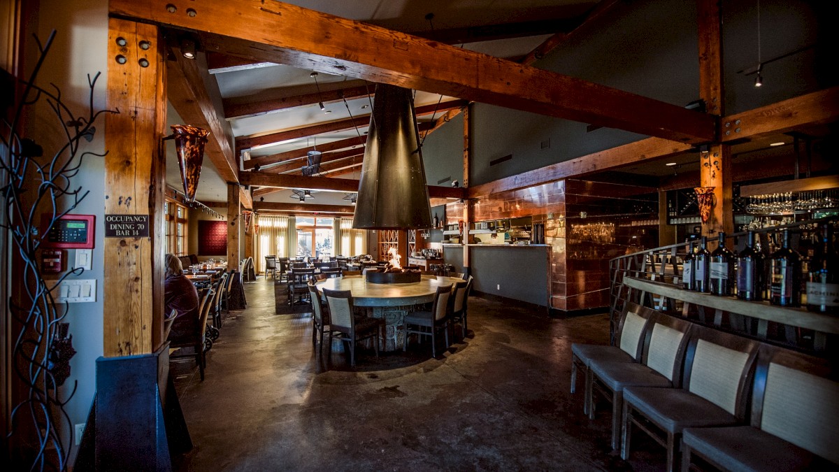 A cozy restaurant interior with wooden beams, round tables, chairs, and a central fireplace, creating an inviting atmosphere.