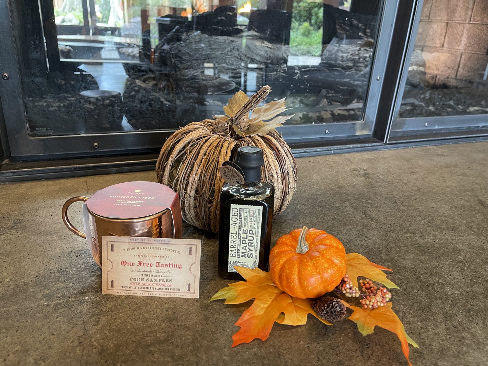 The image shows a fall-themed arrangement with a brown pumpkin, a bottle of olive oil, a mug, and a small orange pumpkin with leaves and pine cones.