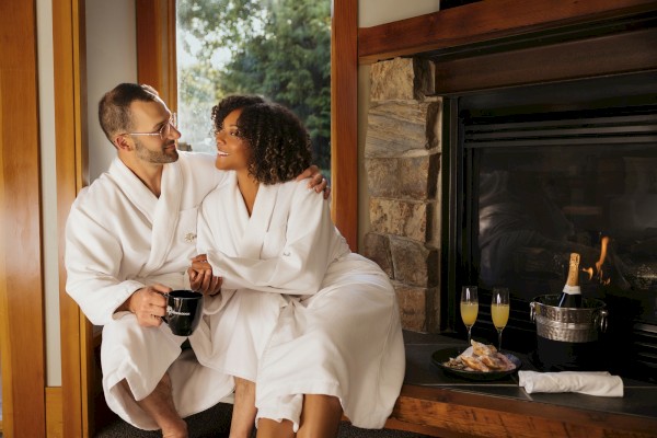 A couple in white robes sits by a fireplace, enjoying drinks and snacks, creating a cozy, intimate setting indoors.