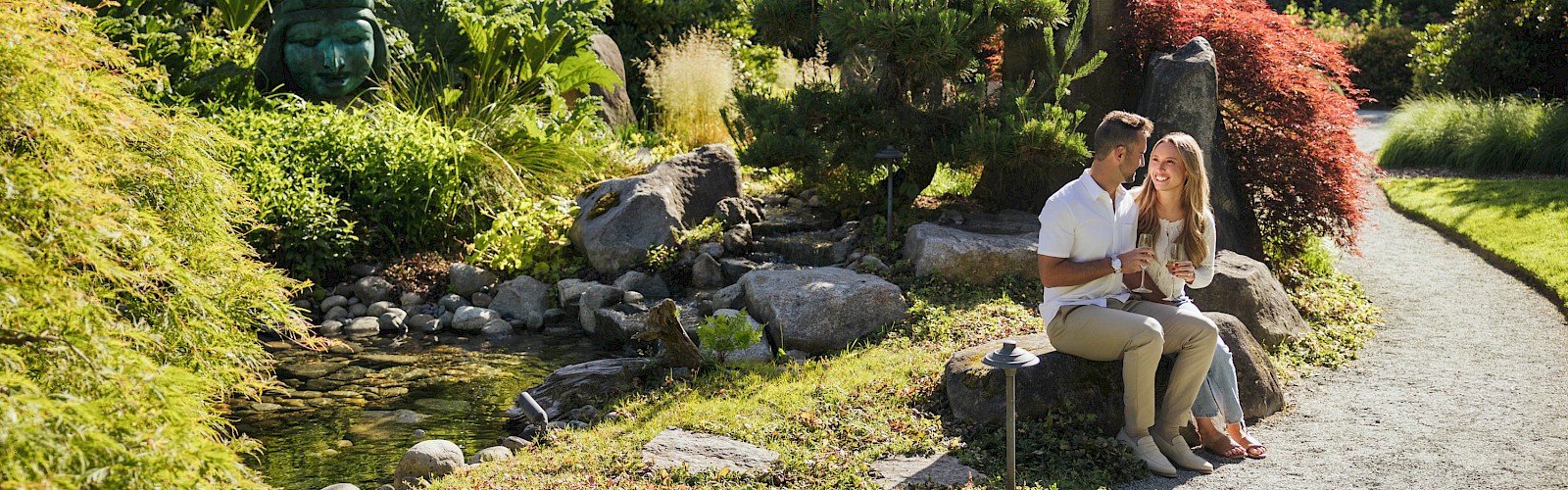 A couple sits on a bench by a rocky garden stream, reading together, with lush greenery and a winding path in a peaceful park setting.