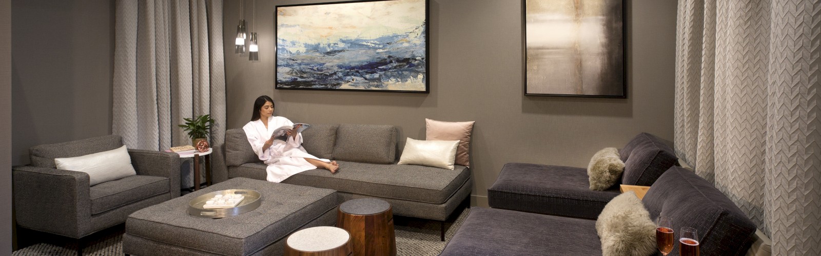 A modern living room with gray sofas, two paintings on the wall, and a person reading. There's a cozy vibe with warm lighting.