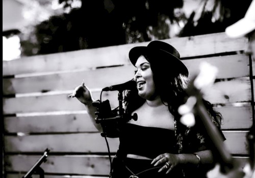 A person singing into a microphone outdoors, wearing a hat and dark clothing, with trees in the background.