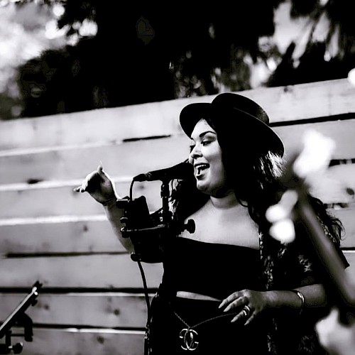 A person singing into a microphone outdoors, wearing a hat and dark clothing, with trees in the background.