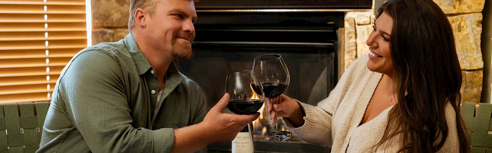 A couple sits by a fireplace, clinking wine glasses and smiling at each other, enjoying a cozy indoor moment.