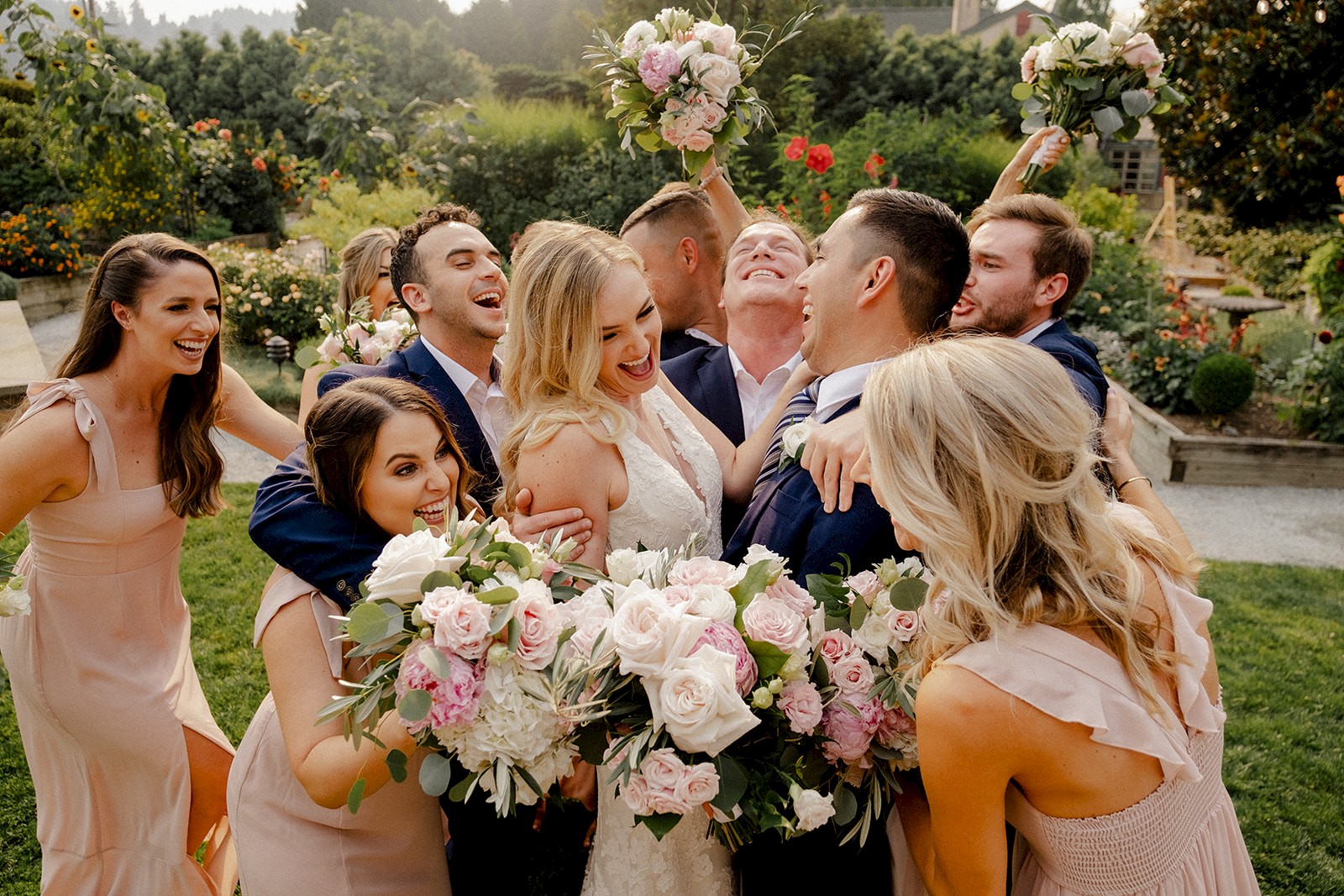 A joyful wedding group photo showing a bride and groom surrounded by smiling friends, all holding bouquets, in an outdoor setting.