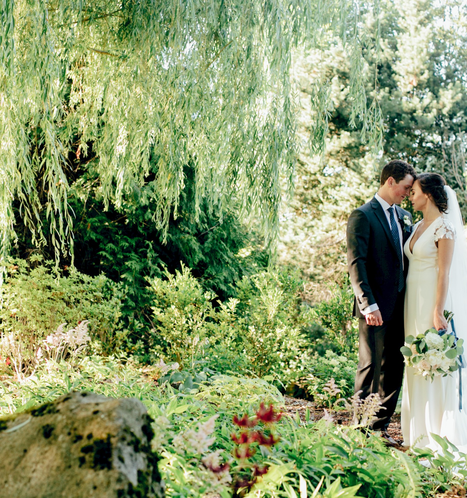 A bride and groom stand in a lush, green garden under a willow tree, embracing and holding a bouquet.
