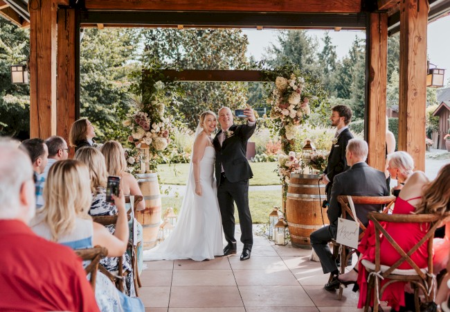 A couple is taking a selfie during an outdoor wedding ceremony with guests seated around, surrounded by floral arrangements and greenery.