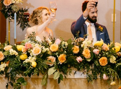 A couple toasts at a floral-decorated table, raising wine glasses. The setting exudes a celebratory, possibly wedding atmosphere.