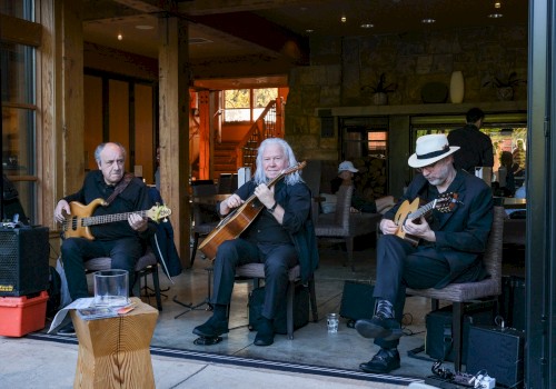A three-member band plays acoustic instruments indoors at a cozy venue with wood accents, while people enjoy a live music performance.