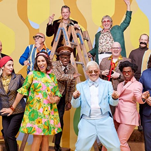 A group of people dressed in colorful retro outfits poses energetically against a vibrant, abstract patterned background.