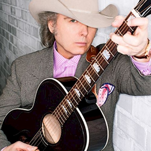 A person in a cowboy hat holds a guitar, wearing a striped shirt and blazer, leaning against a white brick wall.