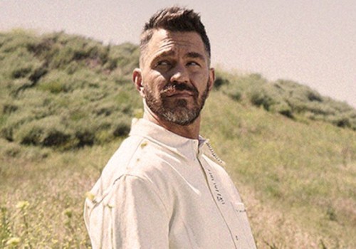 A bearded man in a light-colored shirt stands in a grassy field, looking to the side, with hills in the background.