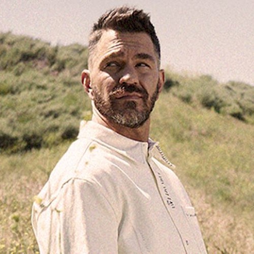 A bearded man in a light-colored shirt stands in a grassy field, looking to the side, with hills in the background.
