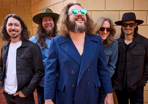 A group of five people standing against a brick wall, wearing casual and stylish outfits with sunglasses and hats, smiling.