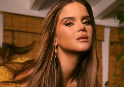 A person with long hair and hoop earrings is posing indoors near wooden blinds and plants.