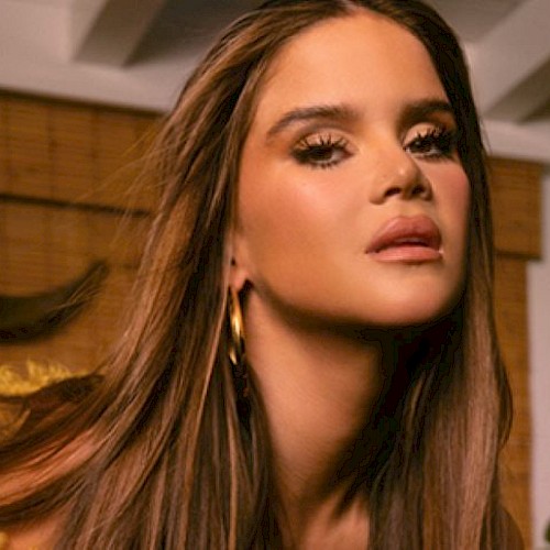 A person with long hair and hoop earrings is posing indoors near wooden blinds and plants.