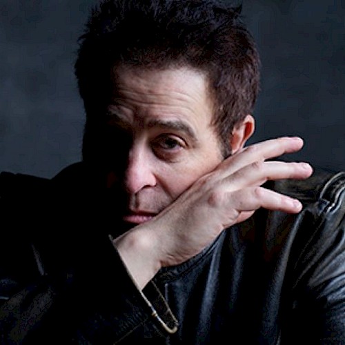 A person in a leather jacket rests their hand near their face, set against a dark background.