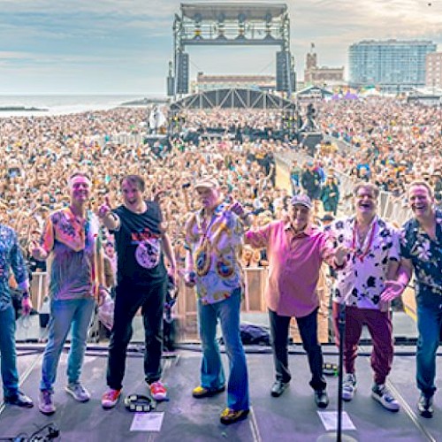 A group of performers on stage poses for a photo with a large crowd in the background at a beachside event, featuring a vibrant atmosphere.