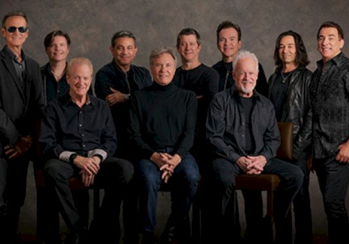 A group of ten people posing together, all dressed in dark clothing, seated and standing against a neutral background.