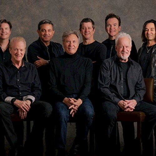 A group of ten people posing together, all dressed in dark clothing, seated and standing against a neutral background.
