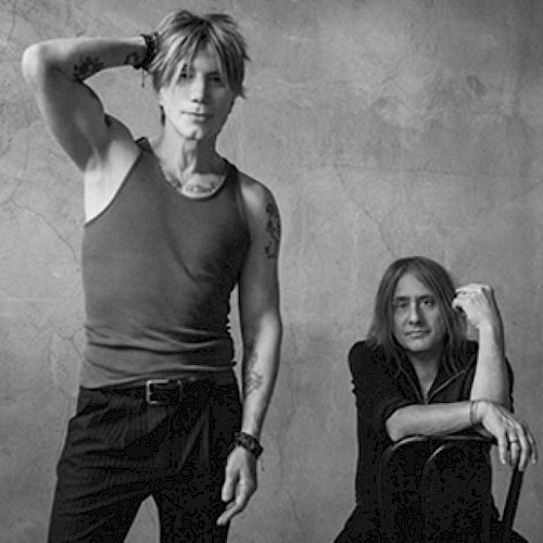 The image shows two people in a black and white photo; one standing with a hand on their head, the other seated, both looking at the camera.