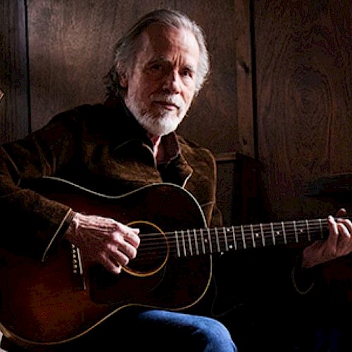 A person with a beard is playing an acoustic guitar, seated in a dimly lit, wooden interior with a decorative emblem on the wall.