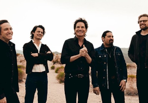 A group of five people standing outdoors in a desert setting, dressed in casual, dark clothing, appear to be enjoying a lighthearted moment.