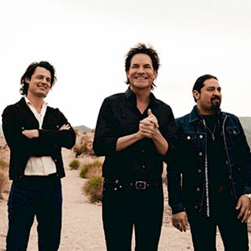 A group of five people standing outdoors in a desert setting, dressed in casual, dark clothing, appear to be enjoying a lighthearted moment.