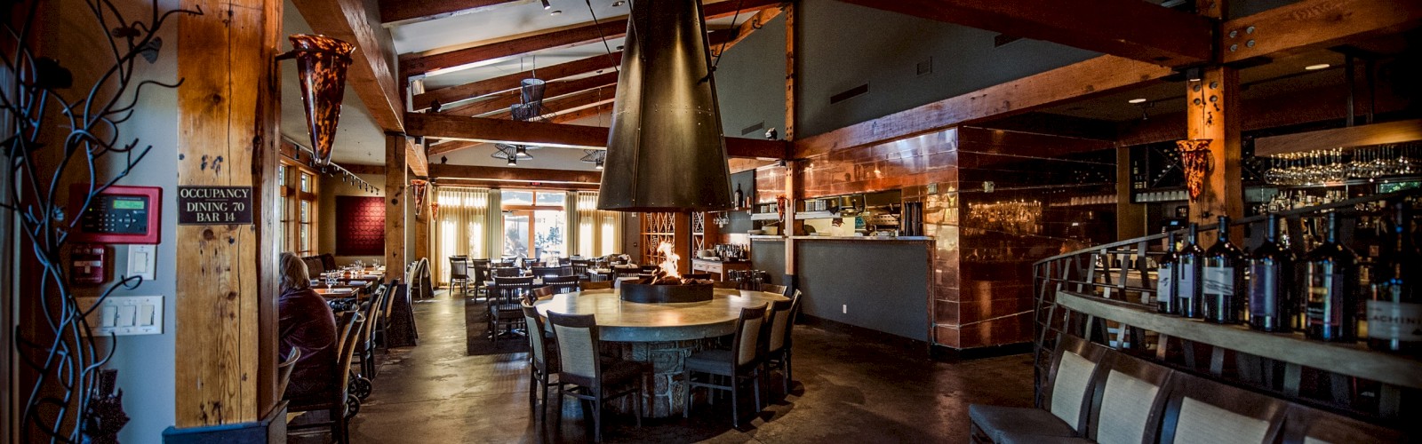 The image shows a modern restaurant interior with wooden beams, tables, chairs, and a bar area, creating a cozy dining atmosphere.