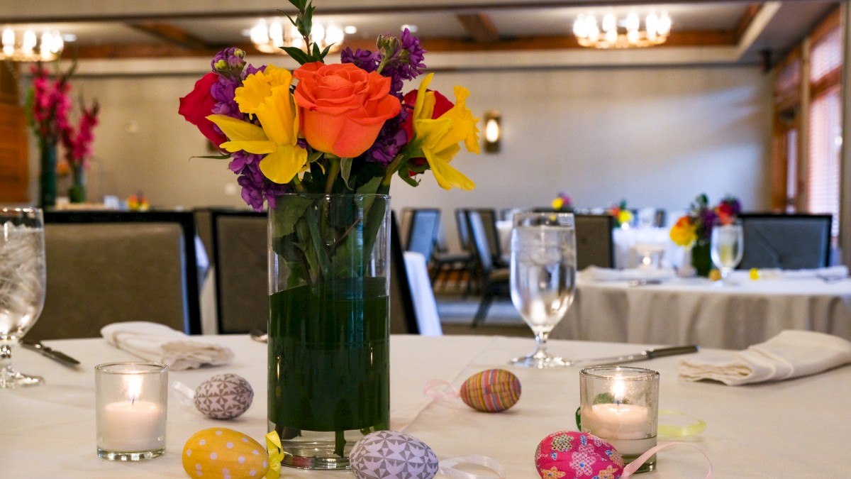 A table decorated with a colorful flower bouquet, lit candles, and decorated eggs for a festive celebration in a dining room.
