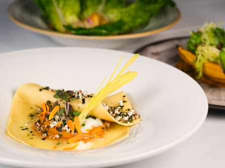 A gourmet dish with a pasta sheet, garnish, and sauce, accompanied by a salad in the background, elegantly plated.