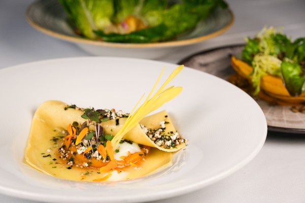 A gourmet dish with a pasta sheet, garnish, and sauce, accompanied by a salad in the background, elegantly plated.