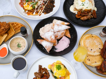 A table with various breakfast dishes, including waffles, pancakes, eggs, fried chicken, and pastries, plus coffee and a drink.