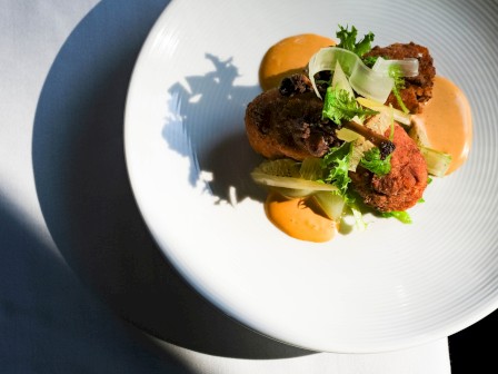 A white plate with gourmet dish featuring crispy items, garnished with greens and sauce, is artfully presented on a white linen.