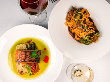 Two plated dishes: one with seared fish in green herb sauce, the other a pasta dish with broccoli and curls, plus a glass of red and a glass of white, seen from above.