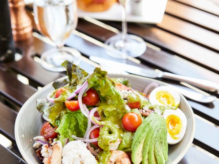 A fresh salad with avocado, eggs, and vegetables is on a table with wine and a sandwich in the background.