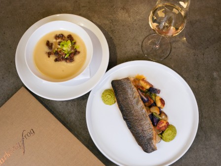 A top-down view of a white bowl of creamy soup with toppings, a plate with a dark eel-like main dish and sides, plus a glass of white wine on a gray tabletop.