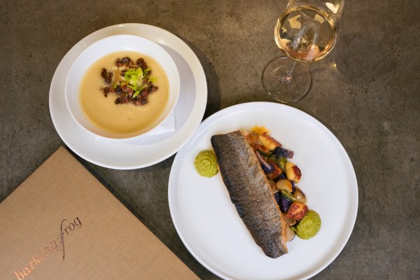 A top-down view of a white bowl of creamy soup with toppings, a plate with a dark eel-like main dish and sides, plus a glass of white wine on a gray tabletop.