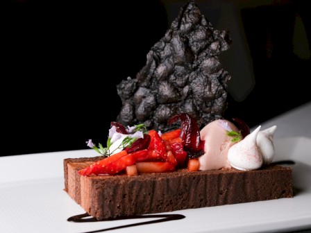 A gourmet chocolate dessert with strawberries, ice cream, mint, edible flowers, and a decorative black chocolate shape on a white plate.