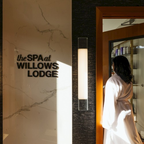 A person in a robe enters a room at the spa at Willows Lodge, with elegant wall lights and a wooden door visible in the setting.