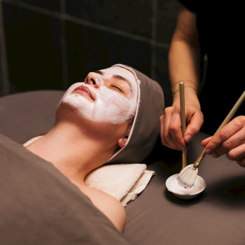 A person is lying on a bed with a white face mask while someone applies it using brushes in a spa setting.