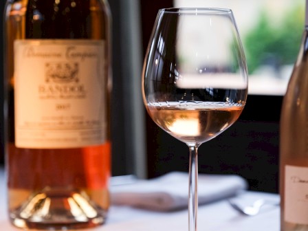 A glass of rosé wine is on a table, with two bottles of wine in the background.
