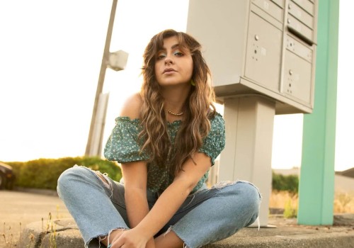 A person with long hair sits on the ground wearing casual clothes and neon shoes, near a mailbox structure on a sunny day.
