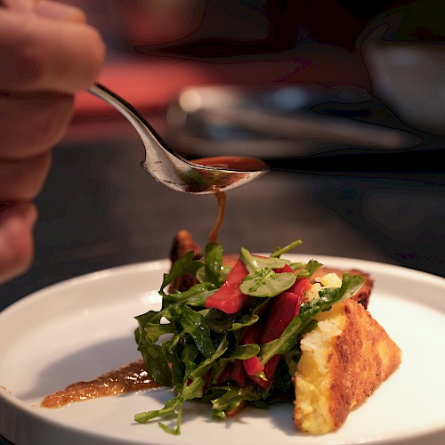 A dish being garnished with dressing, featuring greens and what appears to be a slice of bread or baked item, on a white plate.