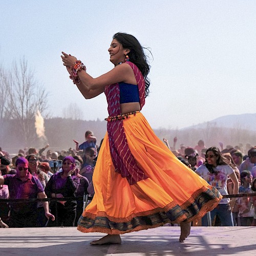 A woman in vibrant traditional attire dances on stage, with a lively crowd celebrating Holi in the background.